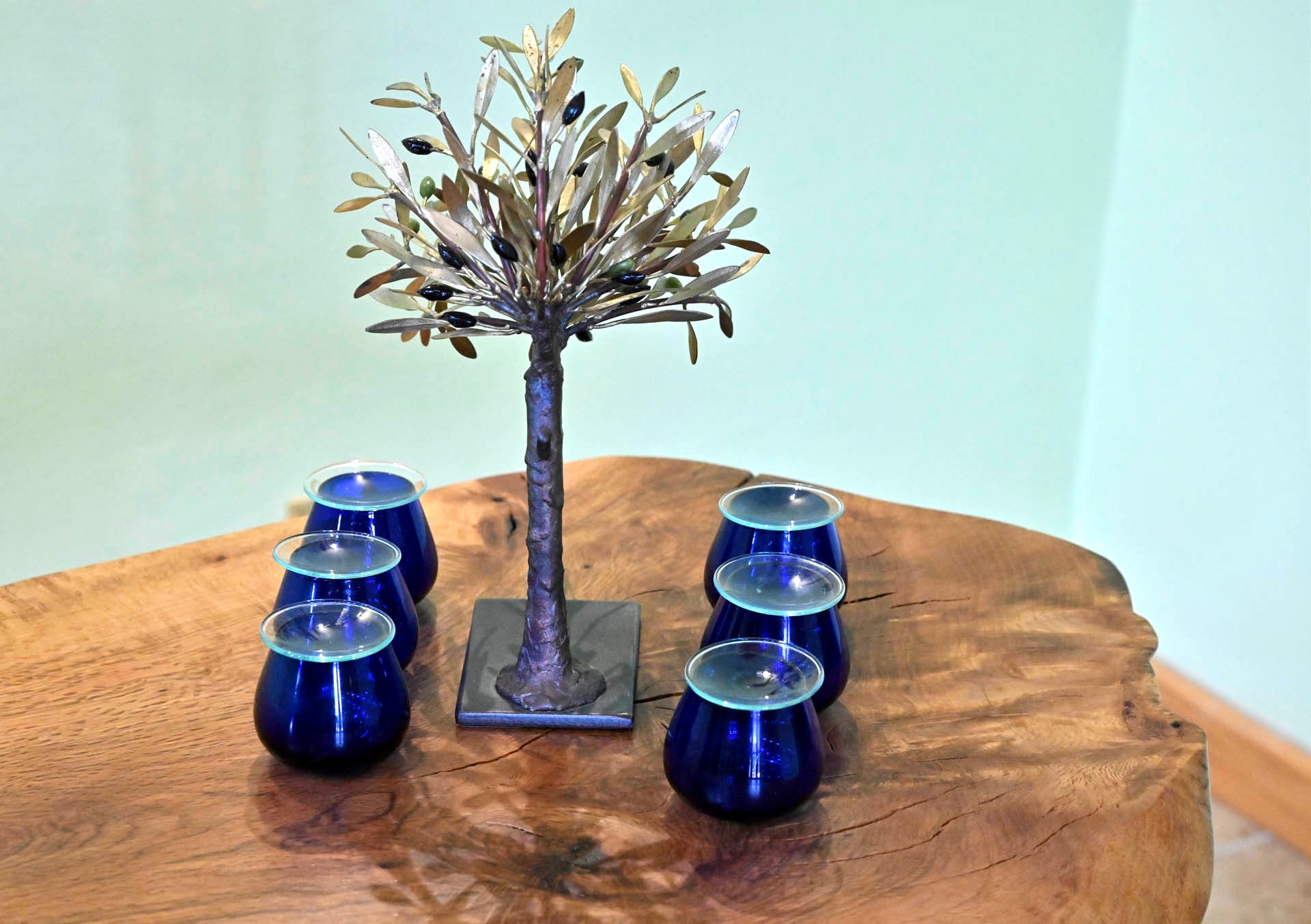Blue olive-oil tasting glasses around a metal olive tree, on an olive-wood table