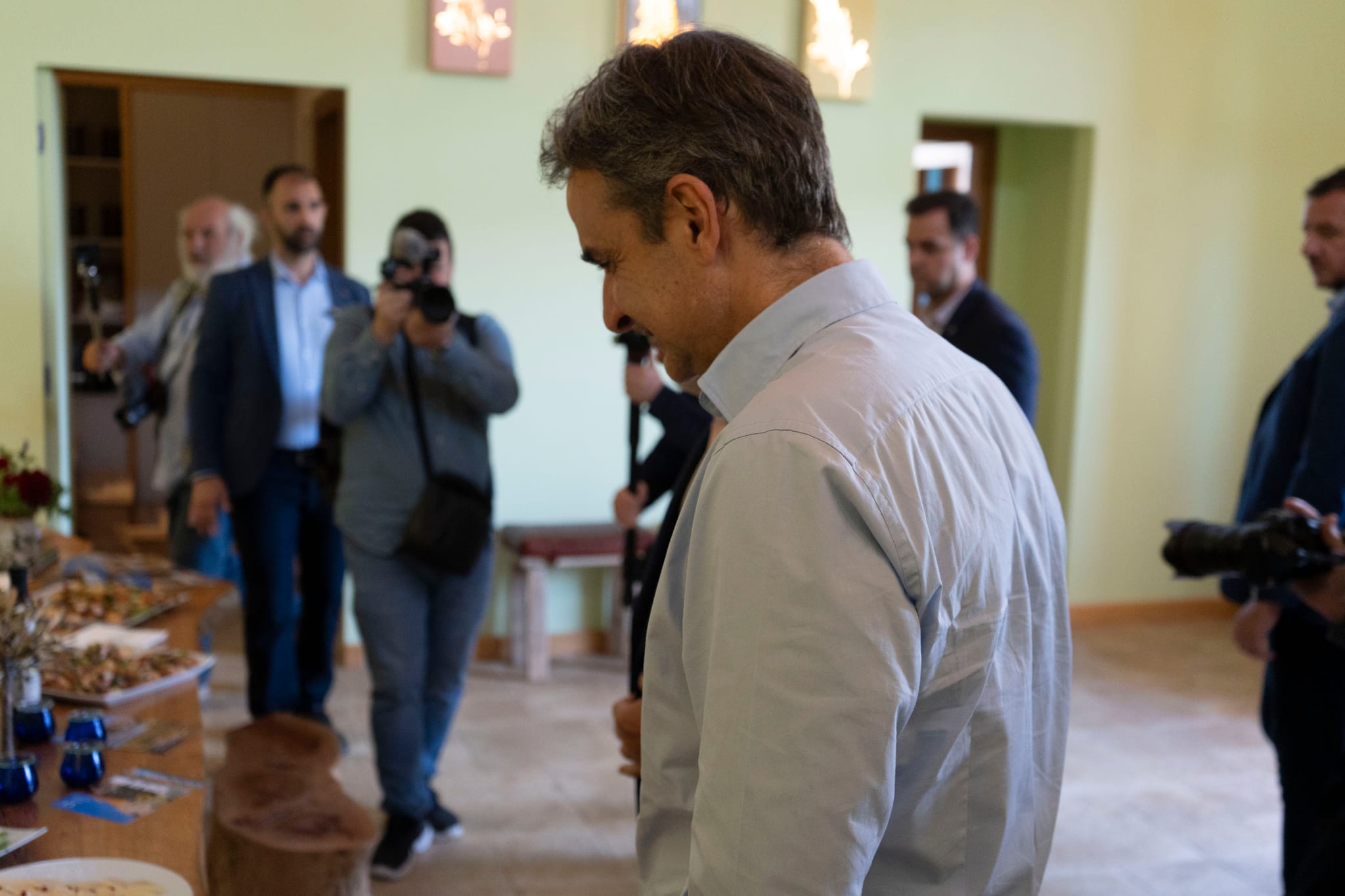 Prime Minister Mitsotakis at a table of local food from Sigri and blue olive oil tasting glasses, as photographers follow